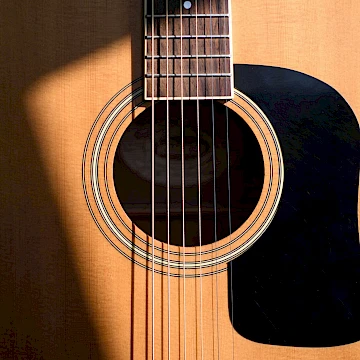 A close up photo of a guitar body, focusing on the opening with the 6 guitar strings running across it. There is a shadow cast across the left side of the guitar.