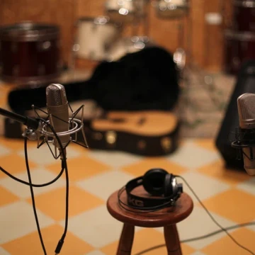 Photo showing a microphone, and headphones on a stool. In the background there is an open guitar case with a guitar in it, a guitar amplifier, and a drum kit.
