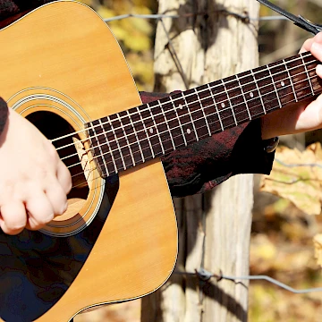 Close up photo of Anya playing a guitar with her hands over the opening and on the top of the neck. In the background is a tree and leaves, the photo was taken outdoors.
