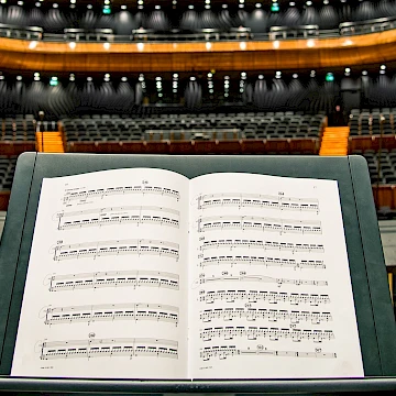 Sheet music on a stand, in the background is an auditorium with black seats and a balcony. The auditorium is constructed with brightly-coloured wood.